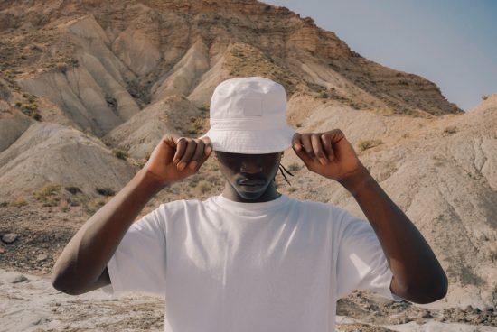 Man in white bucket hat and T-shirt posing in arid landscape, ideal for fashion mockups or diverse model graphics.